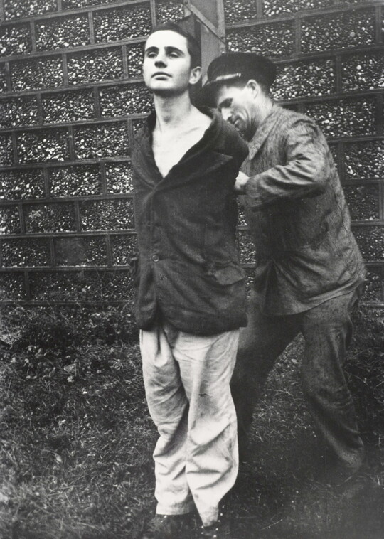 A black-and-white photograph of a young, White man standing with his hands behind his back as another White man ties them to a wood pole.