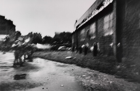 A blurry black-and-white photograph of soldiers on the left shooting rifles at a line of people standing against a wall on the right.