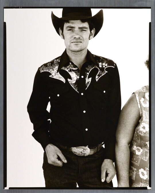 A black-and-white portrait photograph of a White man wearing a dark button-down shirt with Western details, a large belt buckle, and a cowboy hat.