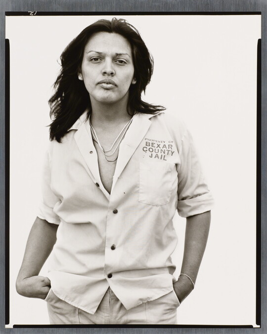 A black-and-white photograph of a medium-skinned person, with long hair wearing a light, partially unbuttoned shirt with text "Property of Bexar County Jail" above the breast pocket.