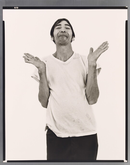 A black-and-white portrait photograph of a medium-skinned man with short hair, light facial hair, wearing a white t-shirt, dark pants, and hands open palmed and raised to shoulders.