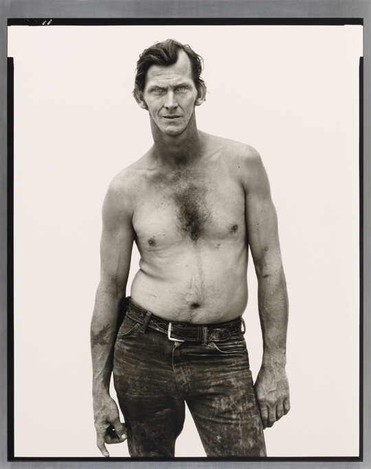 A black-and-white portrait photograph of a shirtless, White man with slicked back hair, wearing distressed jeans, and standing with one hip out.