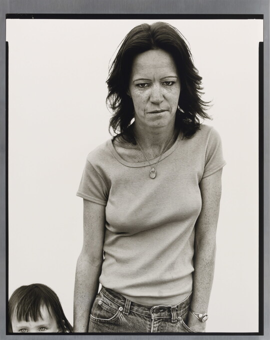 A black-and-white portrait photograph of a weary-looking White woman standing beside a child whose head and body is mostly out of frame.