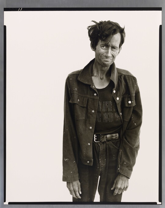 A black-and-white portrait photograph of a thin, tired-looking White man standing in loose-fitting, dark clothes.