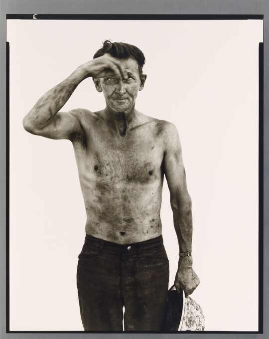 A black-and-white portrait photograph of a dirty, shirtless White man, holding a white hard hat in one hand and shielding his brow with the other.