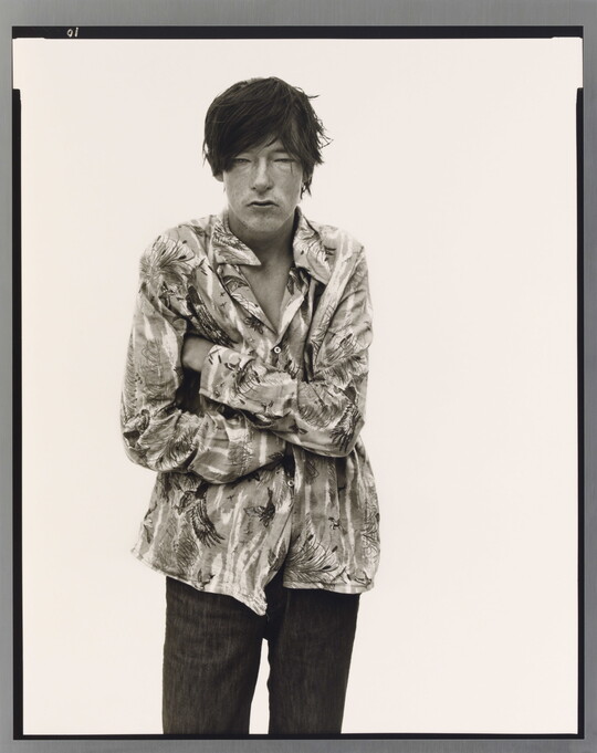 A black-and-white portrait photograph of a light-skinned man with dark hair that hangs into his face standing with arms crossed.
