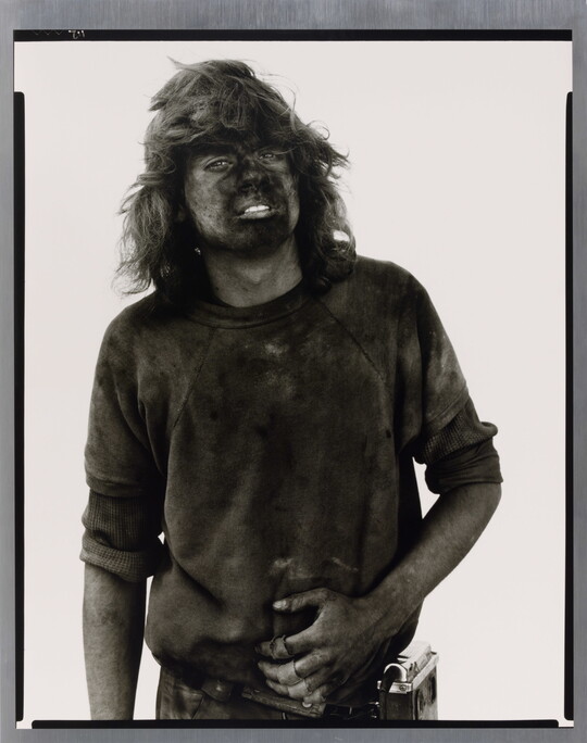 A black-and-white portrait photograph of a young man with long hair covered in dark coal dust.
