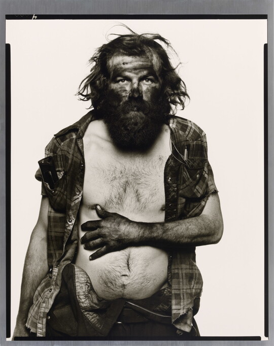 A black-and-white portrait photograph of a bearded man, head and arms covered in dark coal dust, resting his arm across his bare white chest.