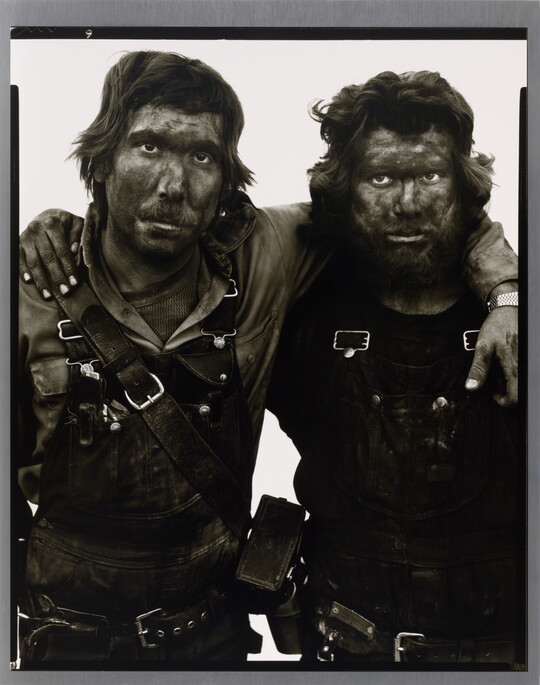 A black-and-white portrait photograph of two men covered in dark coal dust, both wearing dirty overalls, with their arms around one another.