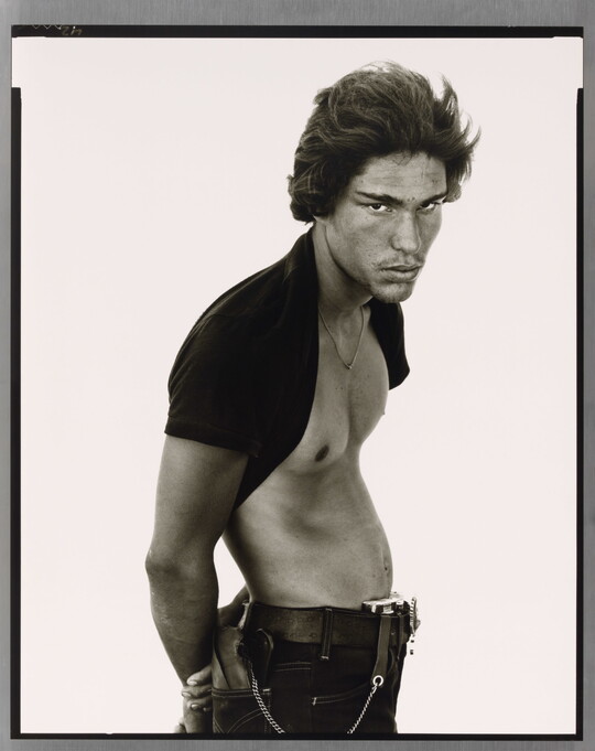 A black-and-white portrait photograph of a medium-skinned man standing sideways, posture in a subtle S-curve, arms behind his back, with a dark t-shirt pulled up behind his neck.