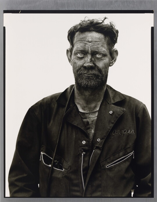 A black-and-white portrait photograph of a man in coveralls, hair sticking up, covered in dark coal dust.