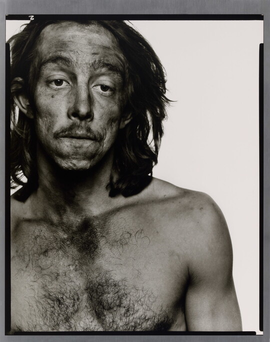A black-and-white portrait photograph of a medium-skinned, shirtless man with long hair, and smudges of dark coal dust on his face.