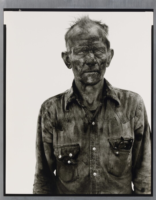 A black-and-white portrait photograph of a man with dirt smudged all over his face and wearing a very dirty button-down shirt.