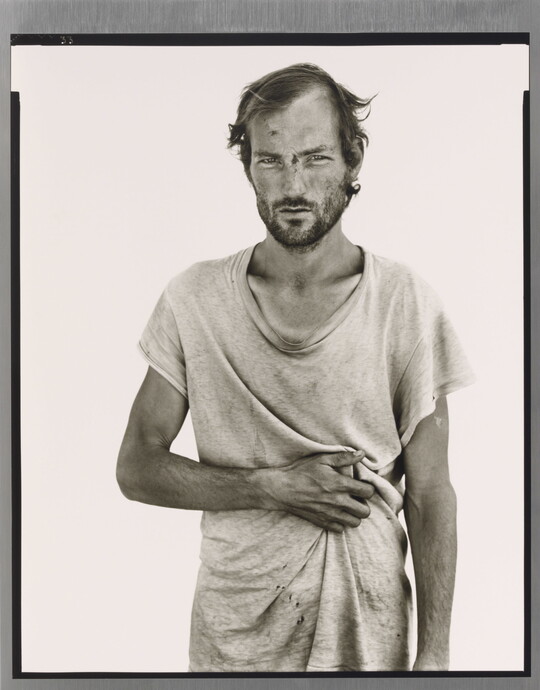 A black-and-white portrait photograph of a White man wearing a loose-fitting dirty white t-shirt, resting his arm across his stomach.