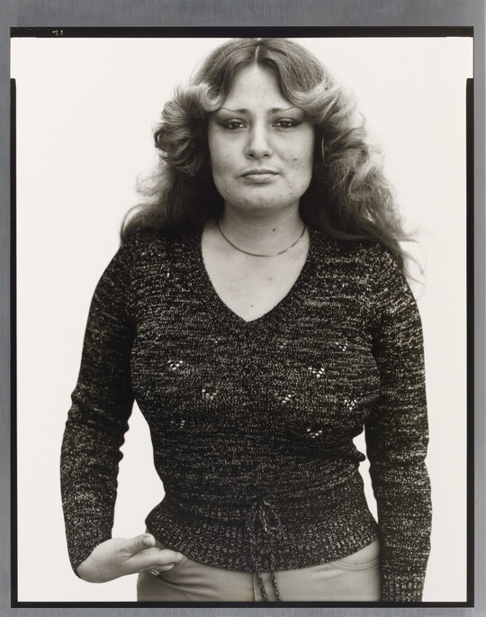 A black-and-white portrait photograph of a White woman with long hair curled back from her face, wearing a fitted dark sweater, and one hand in her pants pocket.