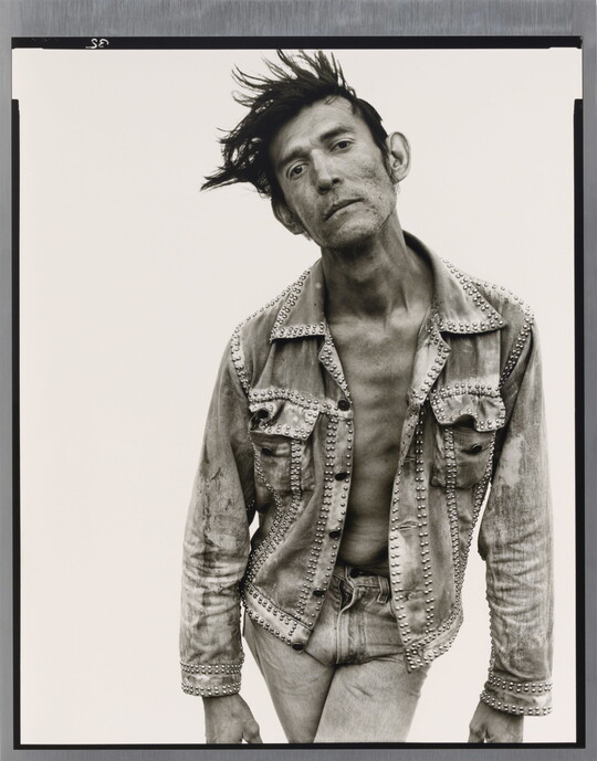 A black-and-white portrait photograph of a White man with dark hair that sticks up, wearing jeans and a jean jacket, and crossing his legs while bending forward.