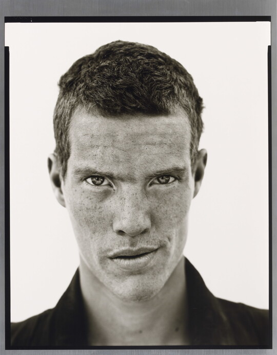 A black-and-white close-up photograph of a White man with short cropped hair, freckles across his face, wearing a dark collared shirt.