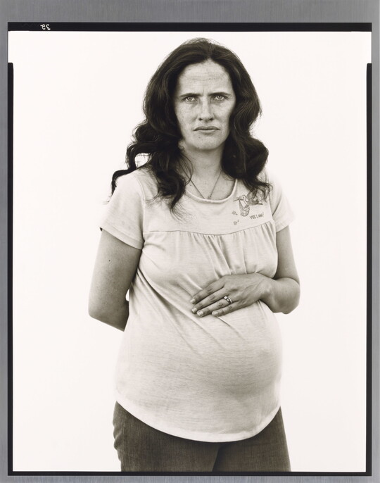 A black-and-white portrait photograph of a pregnant White woman with long, dark hair placing one arm across her stomach and the other behind her back.