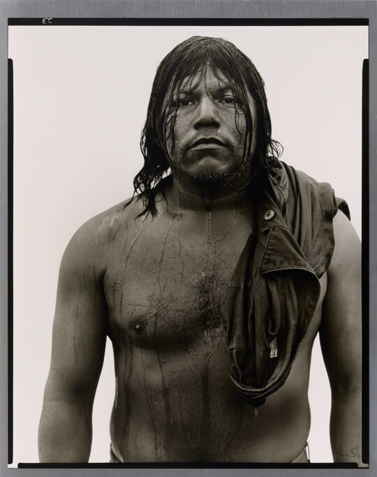 A black-and-white portrait photograph of a stocky, dark-skinned man with long, dark wet hair that drips down his shirtless torso.