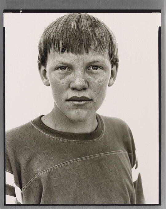 A black-and-white portrait photograph of a White teenage boy with a bowl haircut and freckles across his nose and cheeks, wearing an athletic t-shirt.