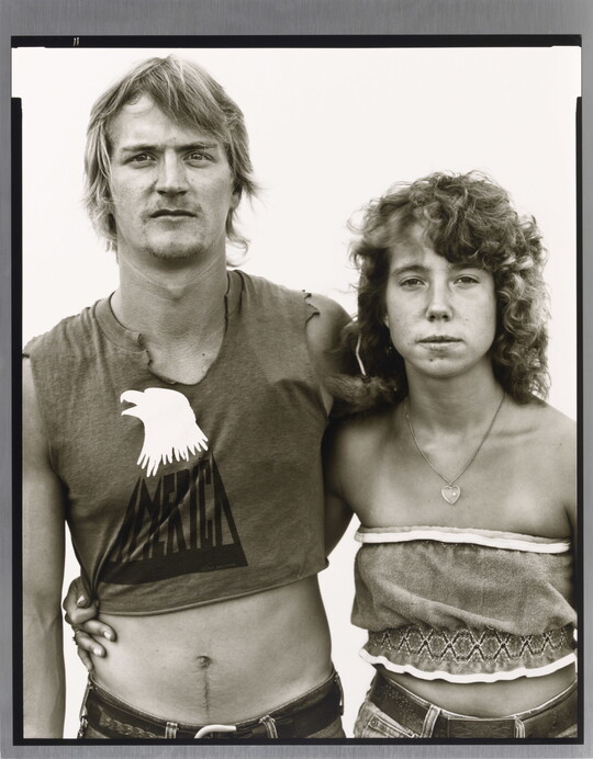 A black-and-white portrait photograph of a young White man and woman, arms around each other, both wearing crop-top shirts.