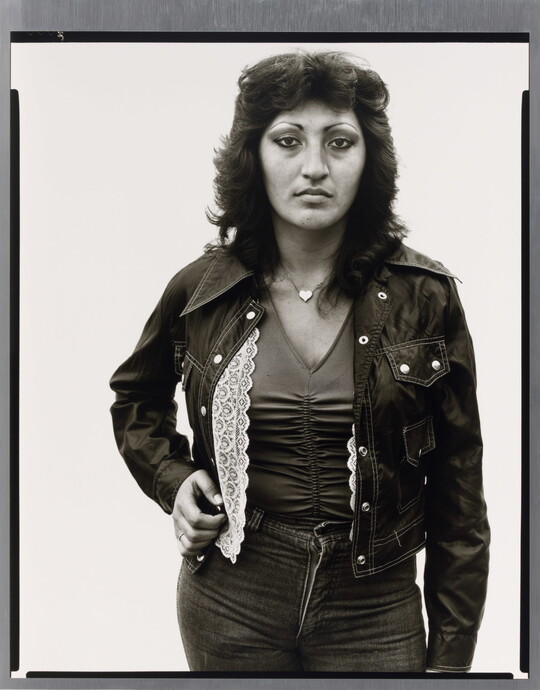 A black-and-white portrait photograph of a medium-skinned woman with dark, feathered hair wearing jeans, a jacket, V-neck top, and a heart necklace.