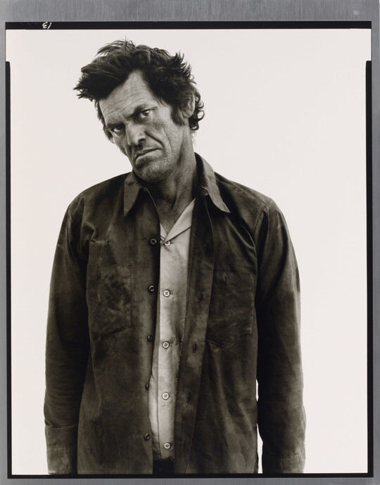 A black-and-white portrait photograph of a White man standing with his head cocked, frowning, and wearing two soiled shirts.