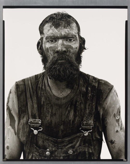 A black-and-white portrait photograph of a bearded man covered in mud and dirt, wearing dirty overalls, and looking straight at the camera.