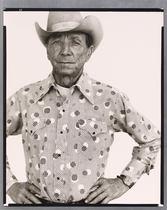 A black-and-white portrait photograph of an older White man wearing a cowboy hat, a button-down shirt, looking straight ahead with hands on hips.