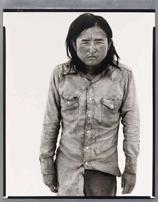 A black-and-white portrait photograph of a medium-skinned man with long hair, wearing dirty clothes, and squinting straight at the camera.