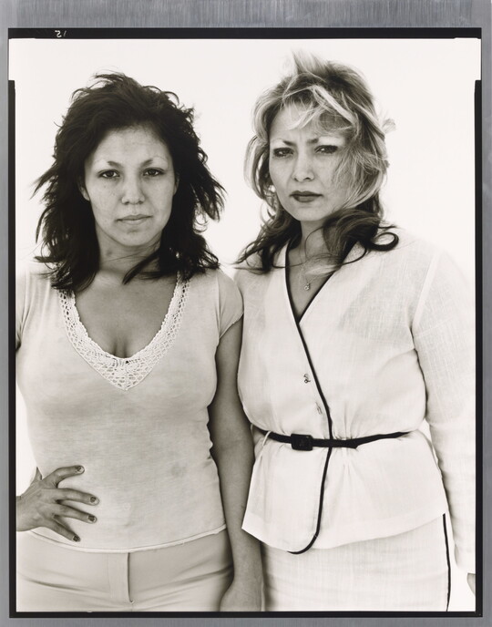 A black-and-white portrait photograph of two medium-skinned women, one in white pants and top with a hand on her hip and the other in a belted white suit, both looking intensely at the camera.