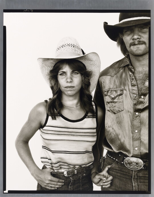 A black-and-white portrait photograph of a young White woman in a cowboy hat and striped tank top with one hand hooked into her jeans and the other hooked into the pocket of a White man in a cowboy hat and satin button-up shirt.