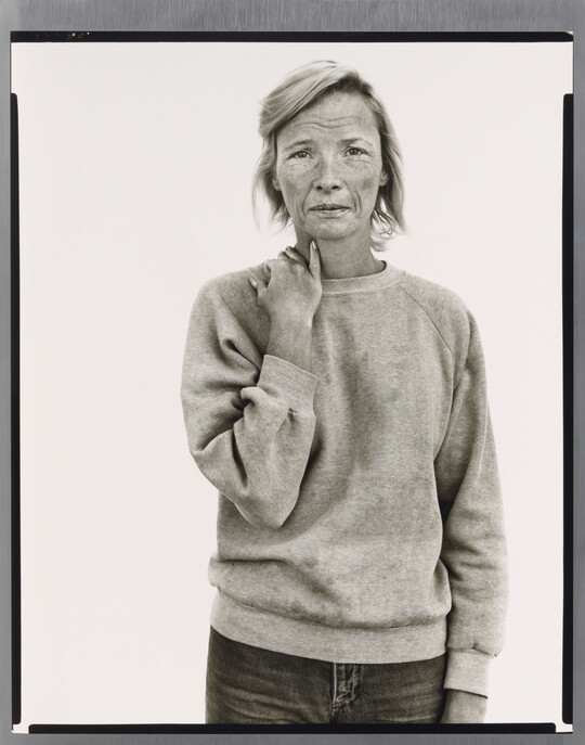 A black-and-white portrait photograph of a tired-looking White woman wearing a sweatshirt and holding her hand on her shoulder as if it might ache.