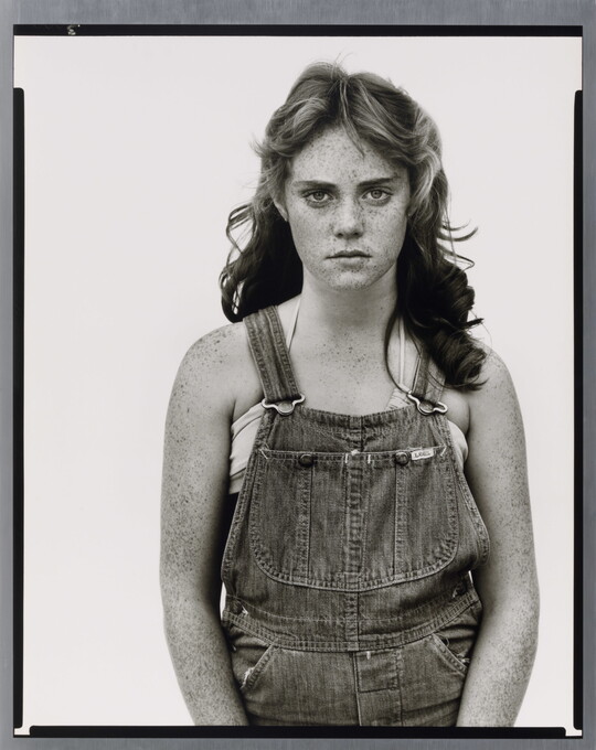 A black-and-white portrait photograph of a young White girl with wavy hair, freckles on her face and arms, wearing denim overalls.