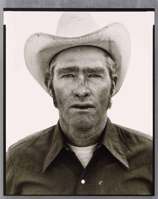 A black-and-white portrait photograph of a White man wearing a cowboy hat and collared shirt, with stubble on his face and chapped lips.