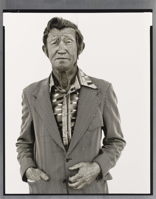 A black-and-white portrait photograph of an older White man with a worn face wearing a patterned, open-collar, button-down shirt under a chevron striped jacket.
