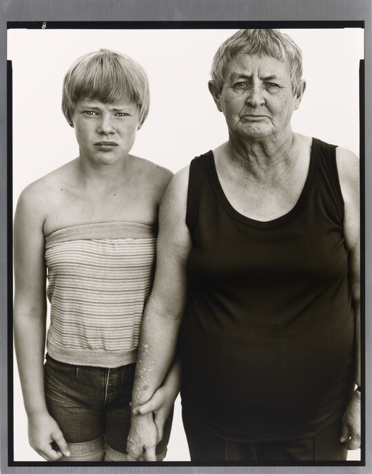 A black-and-white portrait photograph of a young White girl with short blonde hair wearing a tube top, and an older White woman with short gray hair wearing a black tank top.