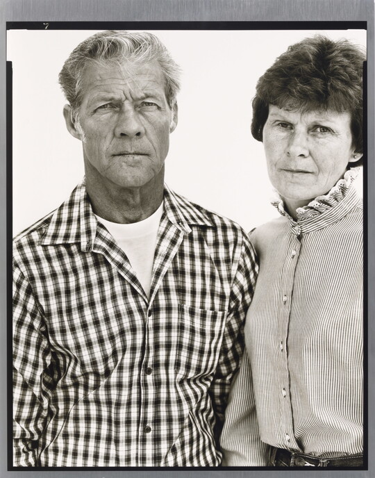 A black-and-white portrait photograph of a middle-aged White man wearing a checked button-down shirt and a middle-aged White woman in a striped high-collar blouse standing shoulder to shoulder.