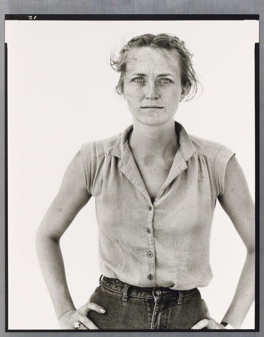 A black-and-white portrait photograph of a young White woman in a sleeveless blouse, hair pulled back from her face, and hands on her hips.