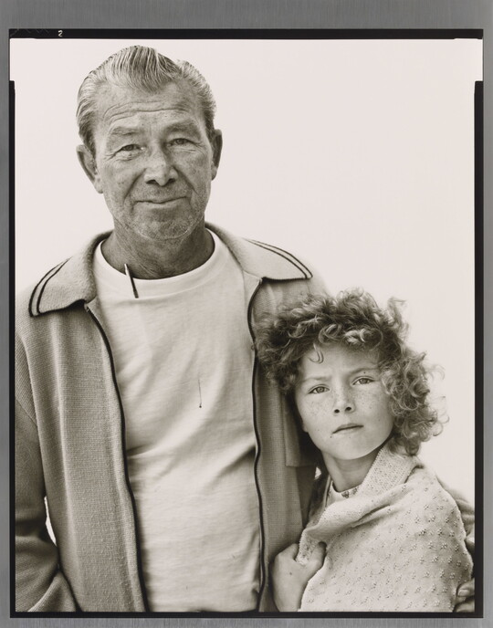 A black-and-white portrait photograph of an older White man in a t-shirt and cardigan with his arm around a young White girl with curly hair and a blanket wrapped around her shoulders.
