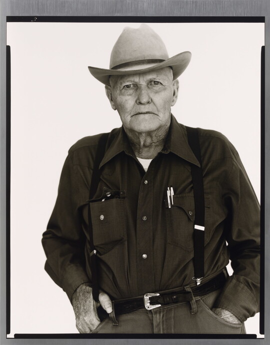 A black-and-white portrait photograph of an older White man wearing a cowboy hat, button down shirt, pants with suspenders and his hands in his pockets