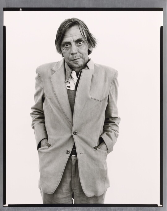 A black-and-white portrait photograph of a middle-aged White man with his hands in the pockets of his unkempt suit jacket.