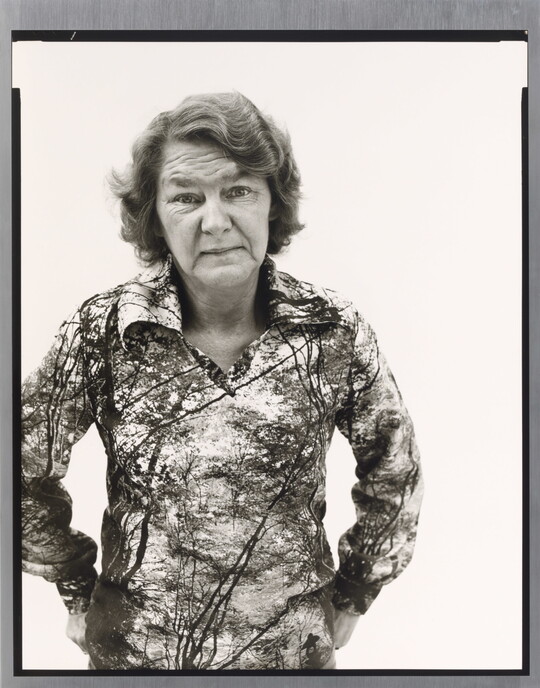 A black-and-white portrait photograph of an older White woman with wavy hair wearing a patterned polo shirt, hands hooked into her back pants pockets.