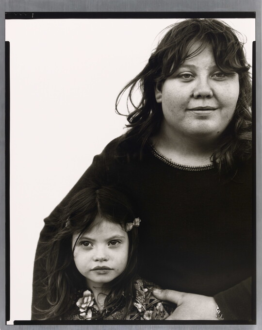 A black-and-white portrait photograph of a medium-skinned woman with dark hair holding close to her a young medium-skinned girl with long dark hair in barrettes.