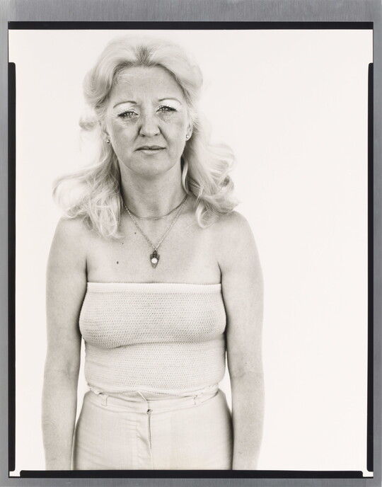 A black-and-white portrait photograph of a White woman with long, bleach-blonde hair in a white tube top and pants.