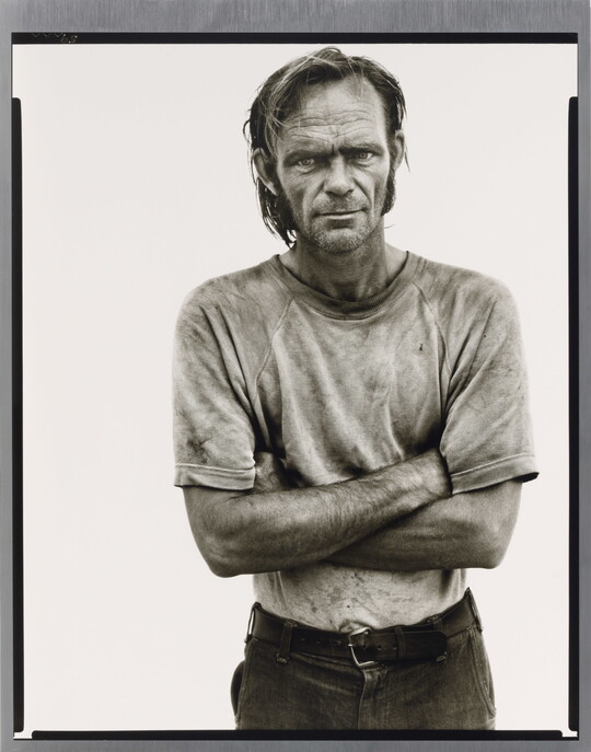 A black-and-white portrait photograph of a White man with unkempt hair in a dirty tee-shirt standing with arms crossed.
