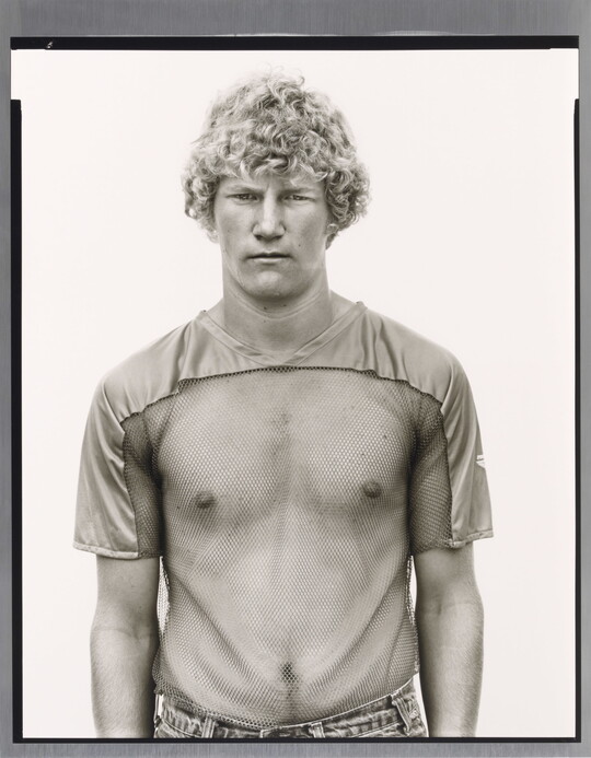 A black-and-white photograph of a young White man with curly blonde hair wearing a see-through, mesh shirt.