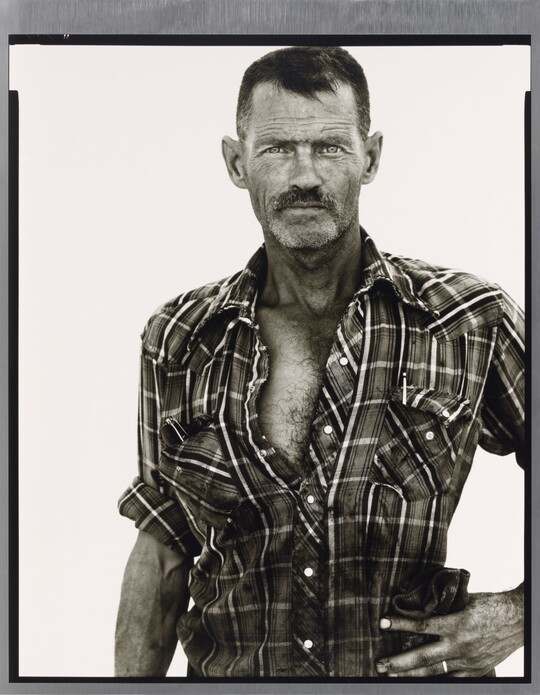 A black-and-white portrait photograph of a middle-aged White man with scruffy facial hair wearing a plaid button-down shirt standing with one hand on his hip.