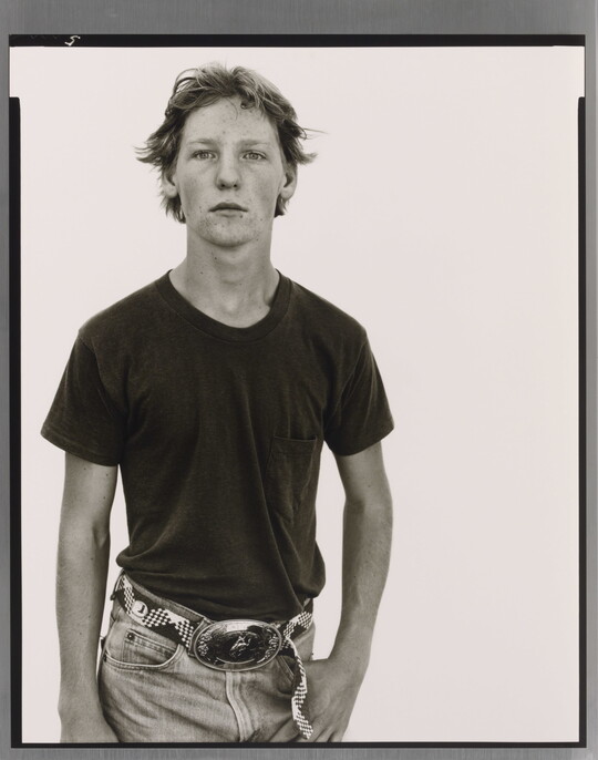 A black-and-white portrait photograph of a White teenage boy in a dark t-shirt and belted jeans with a large belt buckle, one hand hooked into his jeans pocket.