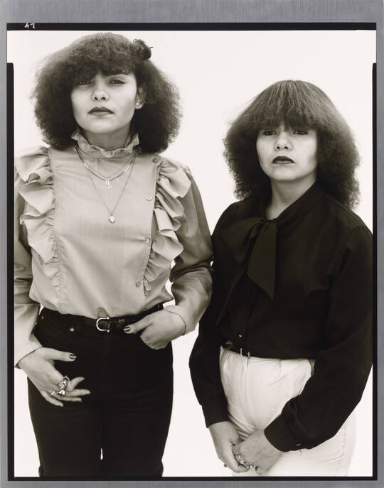 A black-and-white portrait photograph of two medium-skinned young women, both with teased dark hair and wearing ruffled blouses.